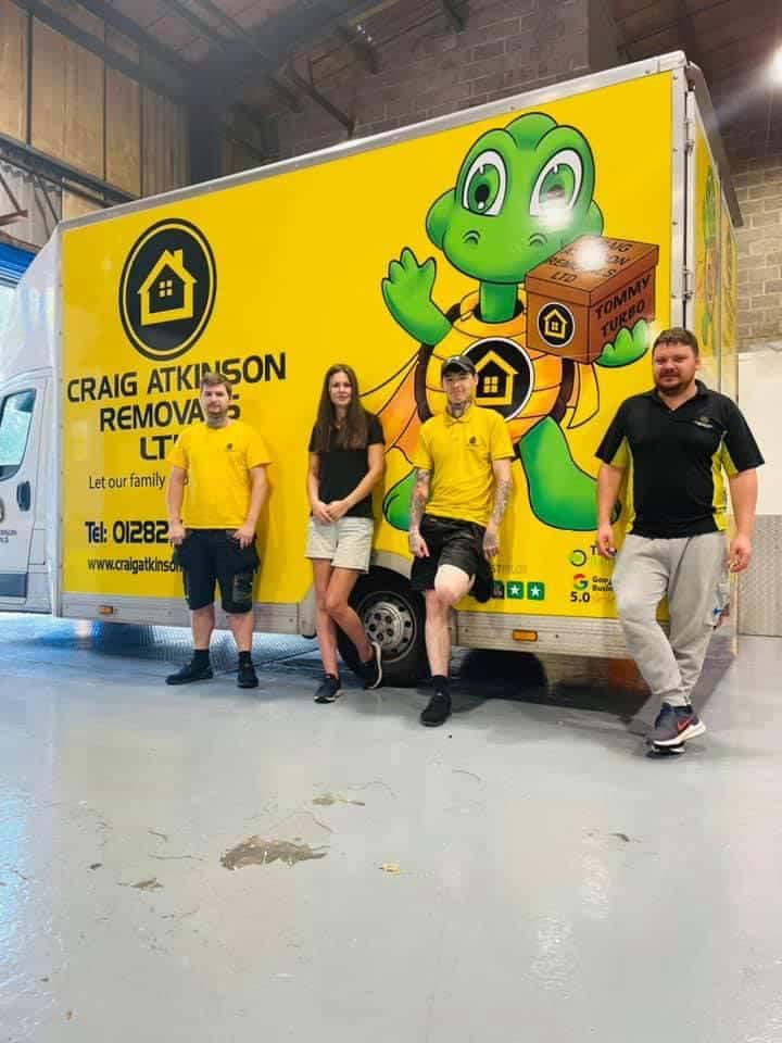 Craig Atkinson Removals team standing beside their bright yellow removals van featuring the Tommy Turbo mascot.