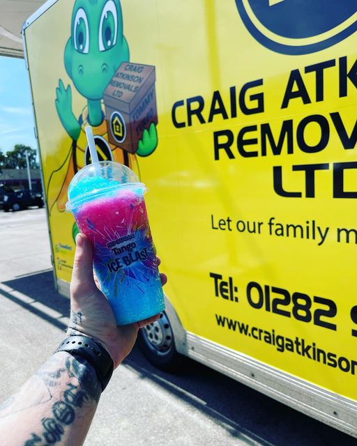 Hand holding a Tango Ice Blast drink in front of a Craig Atkinson Removals van with the turtle mascot.