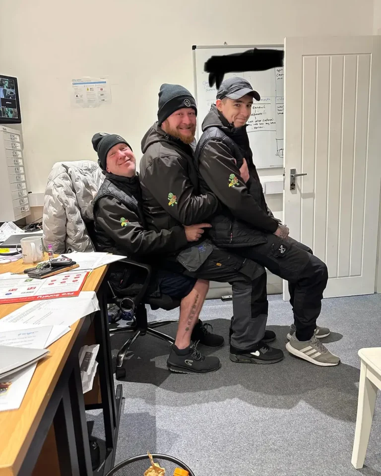 Three Craig Atkinson Removals team members sharing a chair and smiling in the office, dressed in branded winter workwear.